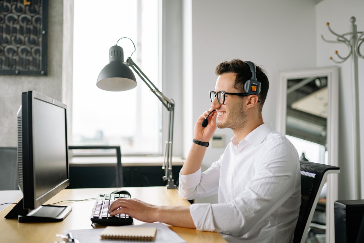 Mastering the First Impression: Your intriguing post title goes here Smiling man wearing headset working at computer in bright office.